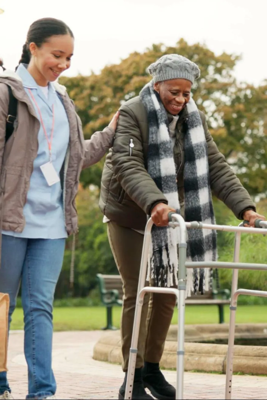 A support worker walking alongside an older woman using a walking frame in a park